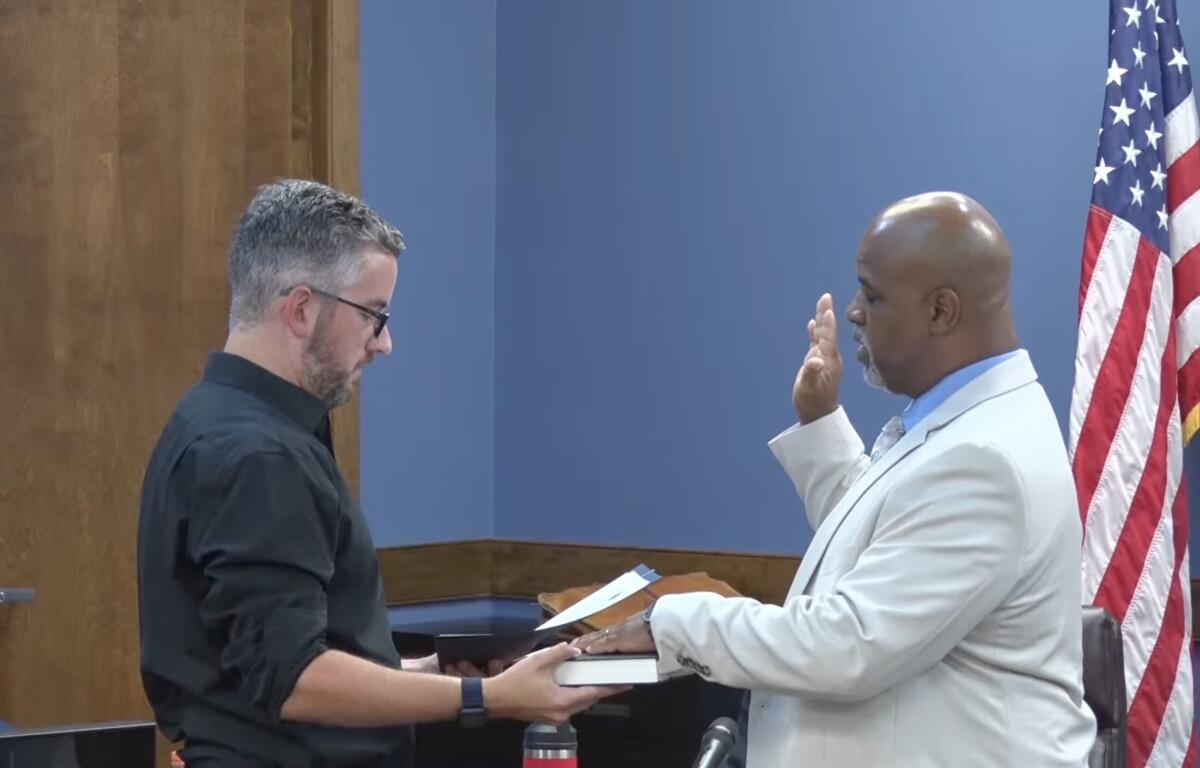 Man in a light suit raises his right hand to take an oath as another man in a black shirt hands him a book, with an American flag in the background.