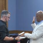 Man in a light suit raises his right hand to take an oath as another man in a black shirt hands him a book, with an American flag in the background.
