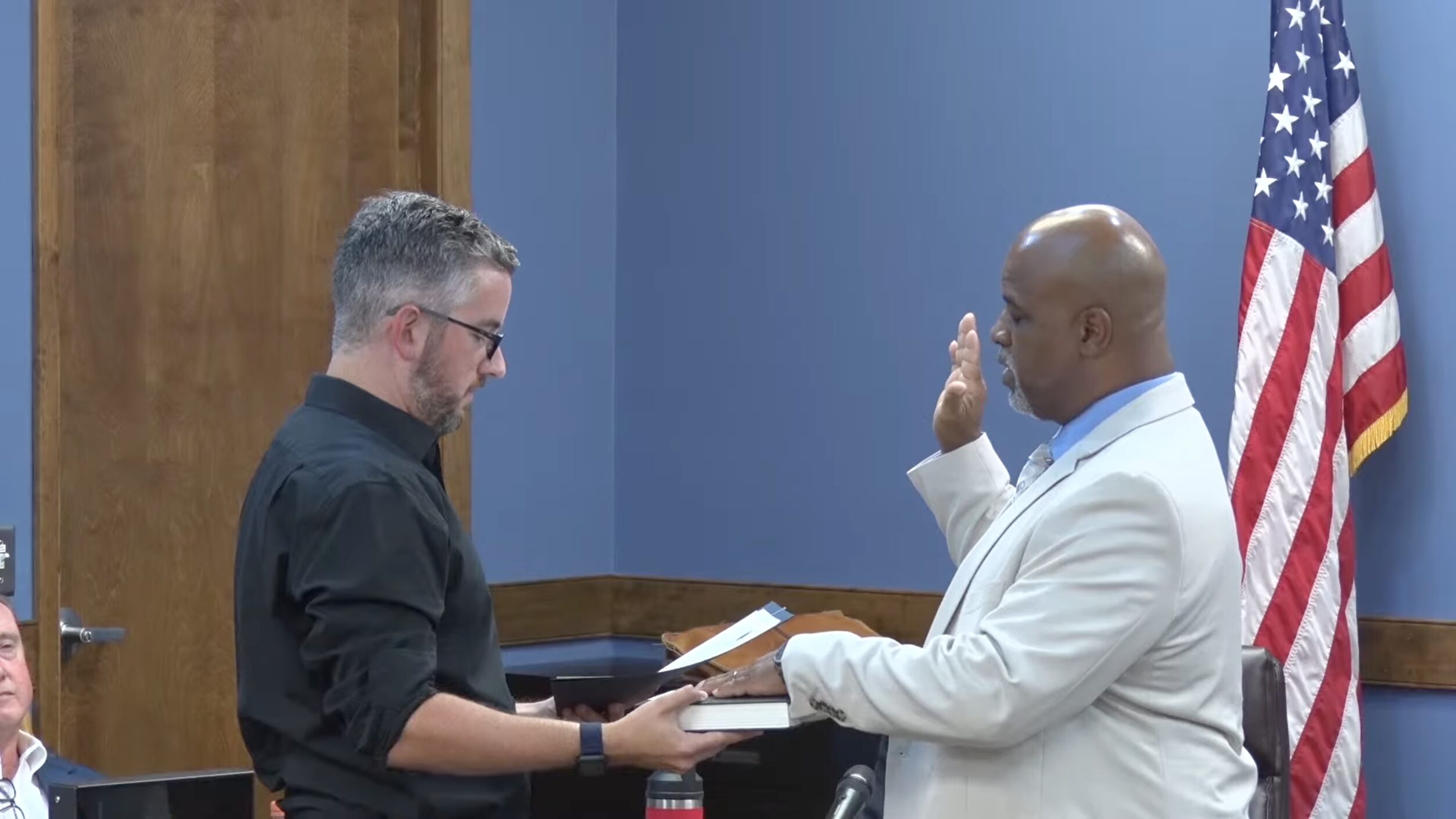 Man in a light suit raises his right hand to take an oath as another man in a black shirt hands him a book, with an American flag in the background.