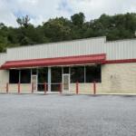 Front view of a small beige storefront with a red metal awning, large glass windows, and a few red bollards in an empty parking lot with trees behind the building.