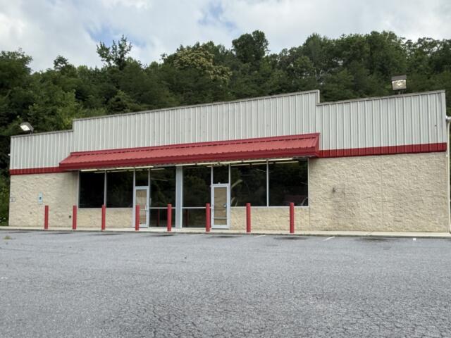 Front view of a small beige storefront with a red metal awning, large glass windows, and a few red bollards in an empty parking lot with trees behind the building.