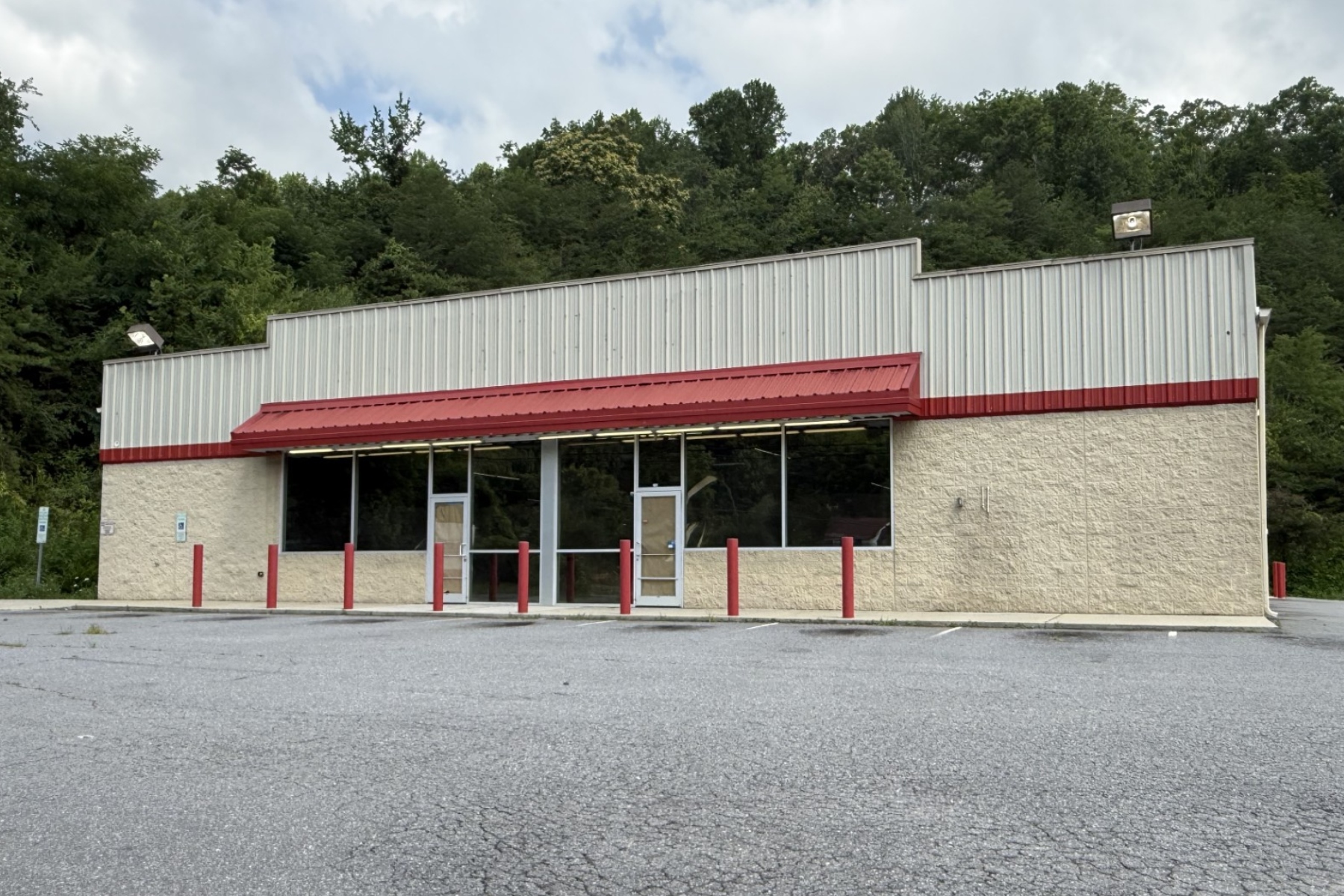 Front view of a small beige storefront with a red metal awning, large glass windows, and a few red bollards in an empty parking lot with trees behind the building.