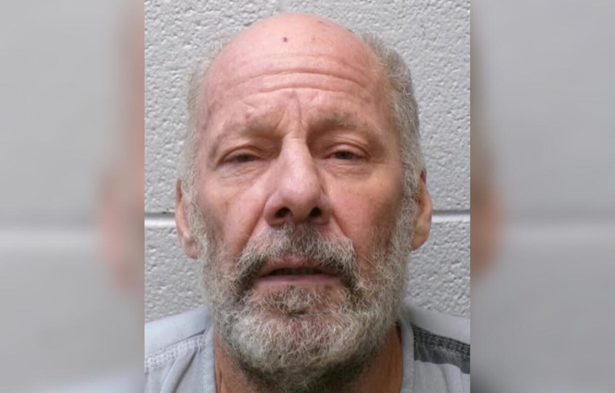 Close-up of an older man with a bald head and gray beard, facing the camera against a gray textured wall.