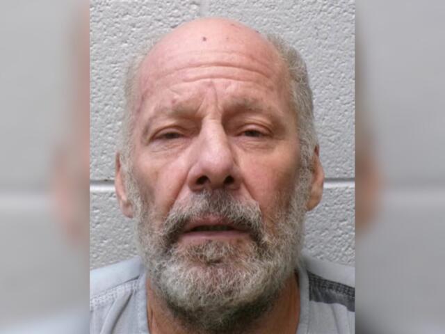 Close-up of an older man with a bald head and gray beard, facing the camera against a gray textured wall.
