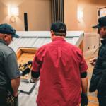 Three men stand around a wooden toolbox, inspecting power tools laid out on a display in a showroom or workshop.