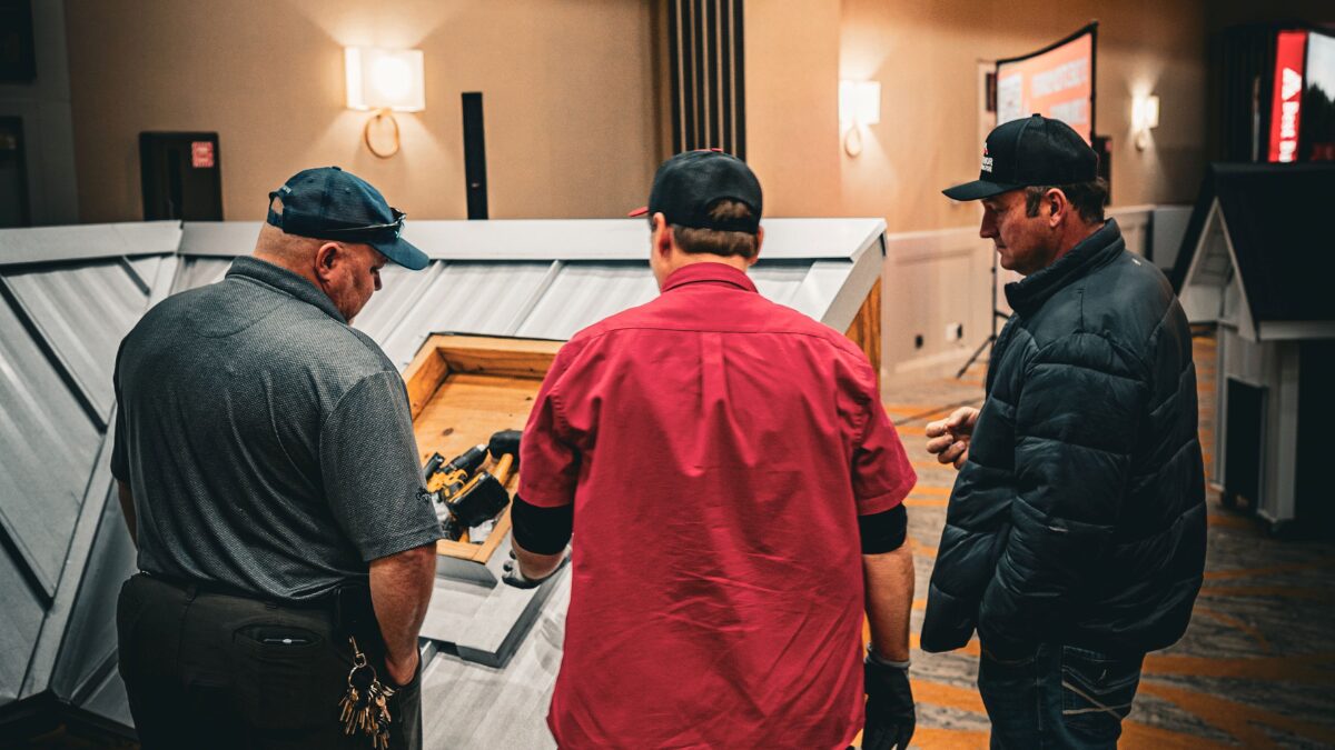 Three men stand around a wooden toolbox, inspecting power tools laid out on a display in a showroom or workshop.