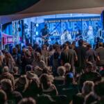 Crowd at an outdoor night concert watching a live band on a blue-lit stage under a canopy.