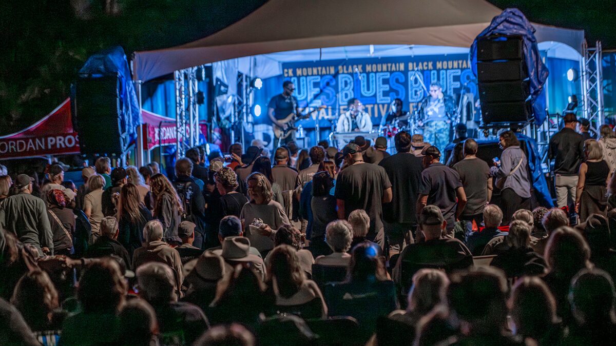 Crowd at an outdoor night concert watching a live band on a blue-lit stage under a canopy.