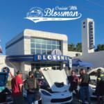 Car show in a sunny lot outside a retro Blossman gas building, with open-hood cars and a crowd of people inspecting them.