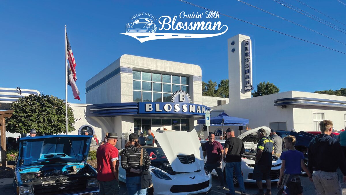 Car show in a sunny lot outside a retro Blossman gas building, with open-hood cars and a crowd of people inspecting them.