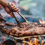 Close-up of a hand using tongs to flip juicy grilled ribs on a barbecue over open flames with smoke rising