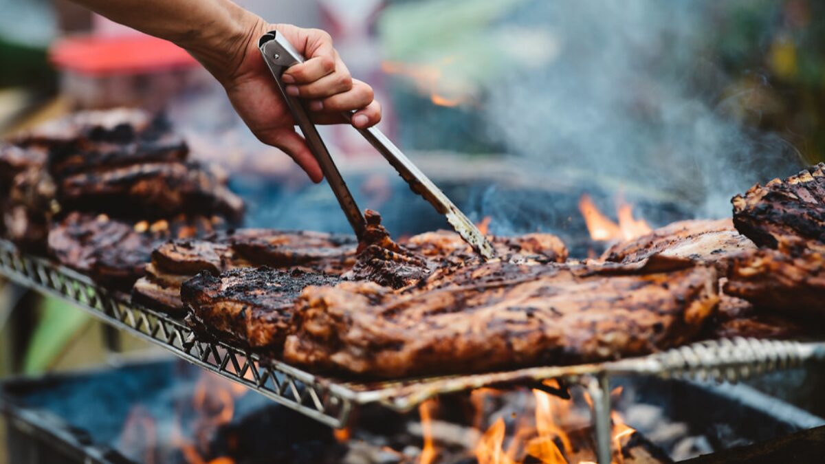 Close-up of a hand using tongs to flip juicy grilled ribs on a barbecue over open flames with smoke rising