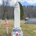 Tall white obelisk tombstone in a cemetery, with flags and flowers at its base and bare trees in the background.