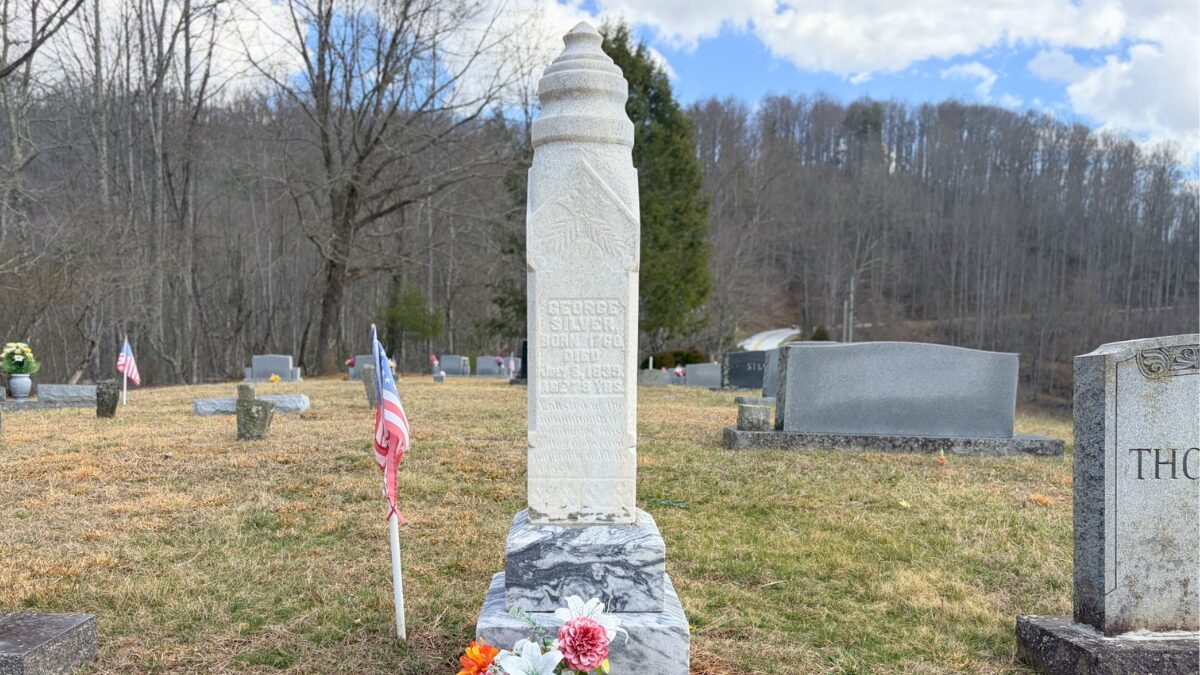 Tall white obelisk tombstone in a cemetery, with flags and flowers at its base and bare trees in the background.