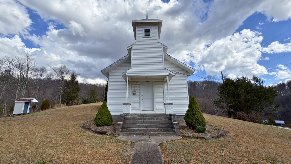 White wooden church on a grassy hillside with stairs leading to the entrance under a partly cloudy sky.