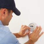 Man in a blue shirt and cap adjusting a round wall thermostat with his hands on a white wall
