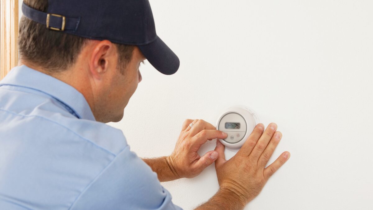 Man in a blue shirt and cap adjusting a round wall thermostat with his hands on a white wall