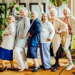 Seven cheerful elderly women pose playfully in a bright, plant-filled room, standing in a line with arms raised.