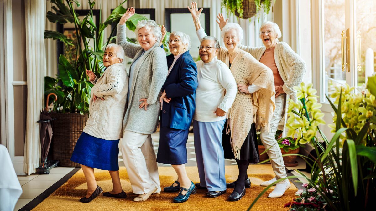 Seven cheerful elderly women pose playfully in a bright, plant-filled room, standing in a line with arms raised.