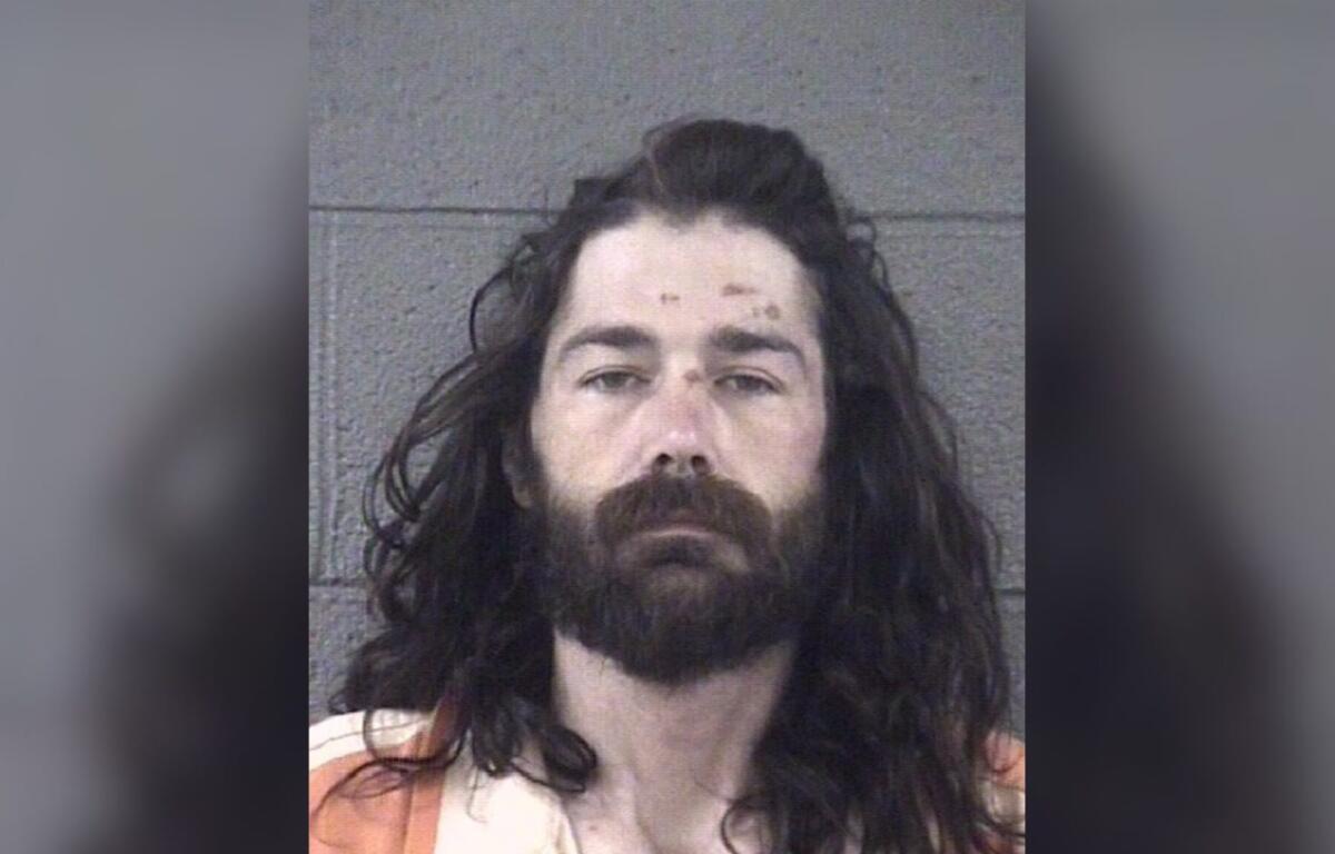 Mugshot of a man with long dark hair and full beard, wearing an orange striped jail uniform in front of a gray wall.