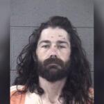 Mugshot of a man with long dark hair and full beard, wearing an orange striped jail uniform in front of a gray wall.