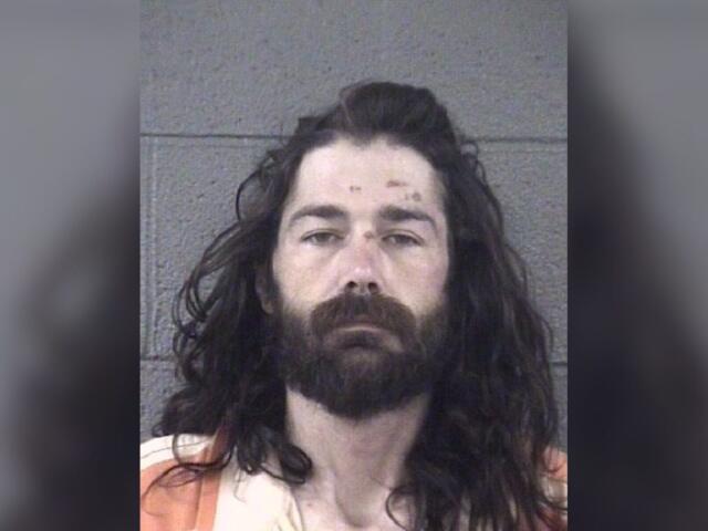Mugshot of a man with long dark hair and full beard, wearing an orange striped jail uniform in front of a gray wall.
