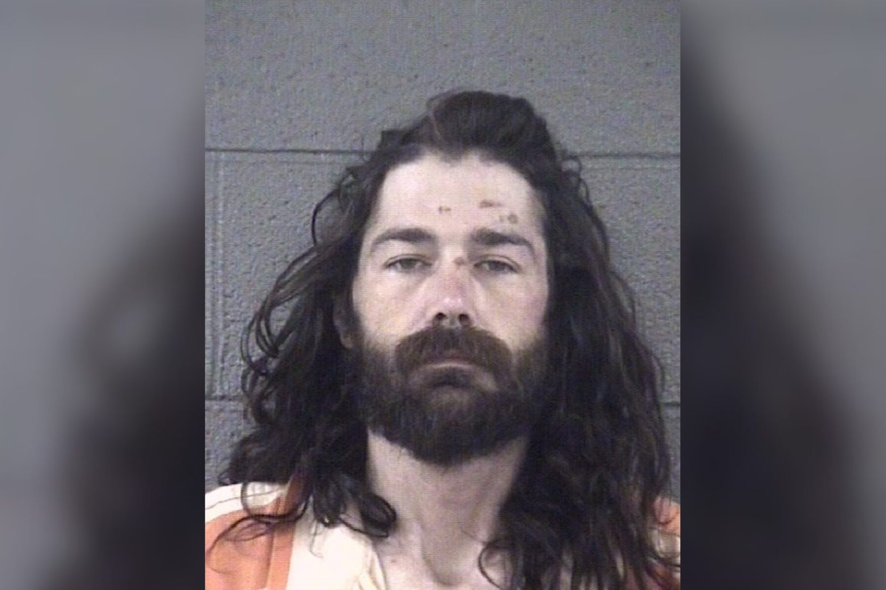 Mugshot of a man with long dark hair and full beard, wearing an orange striped jail uniform in front of a gray wall.
