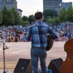 Three musicians with guitars on an outdoor stage facing a large crowd in a city park, with trees and buildings in the background.