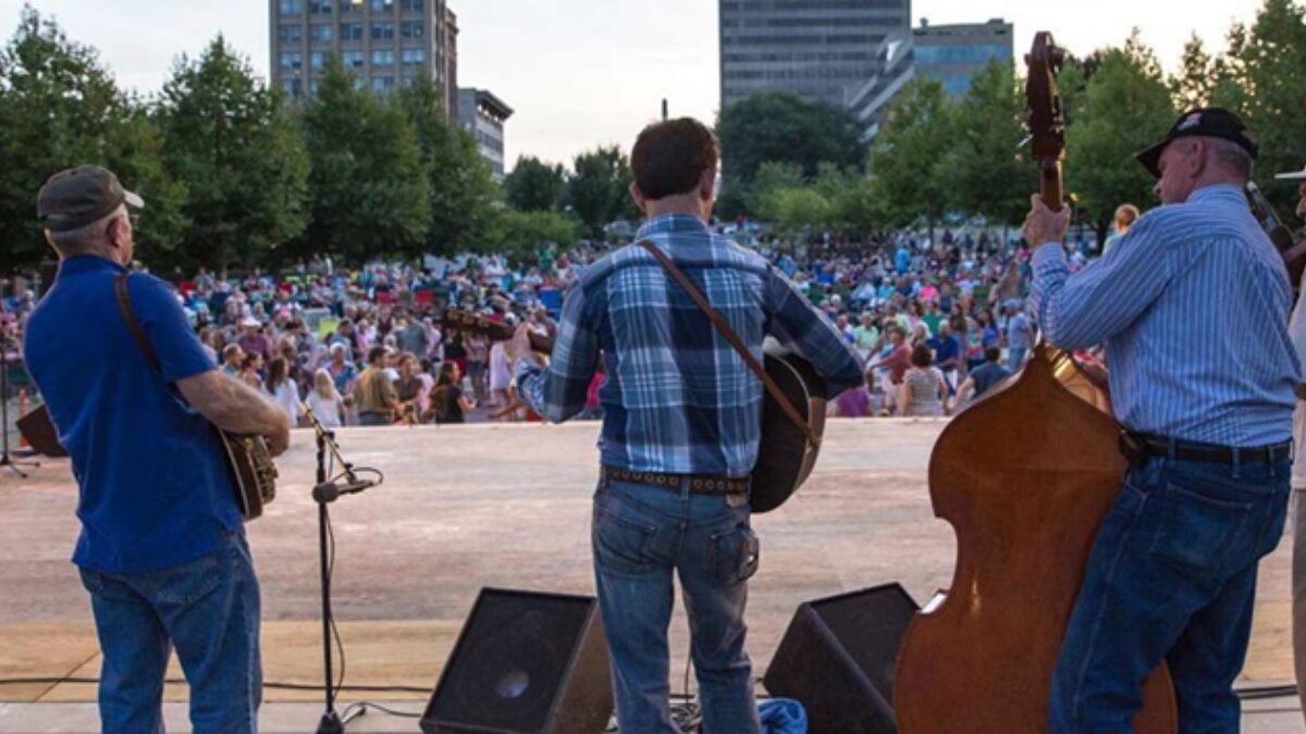 Three musicians with guitars on an outdoor stage facing a large crowd in a city park, with trees and buildings in the background.