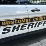 White Buncombe County Sheriff's Office patrol vehicle door with gold and black 'SHERIFF' lettering and sheriff badge visible on the side
