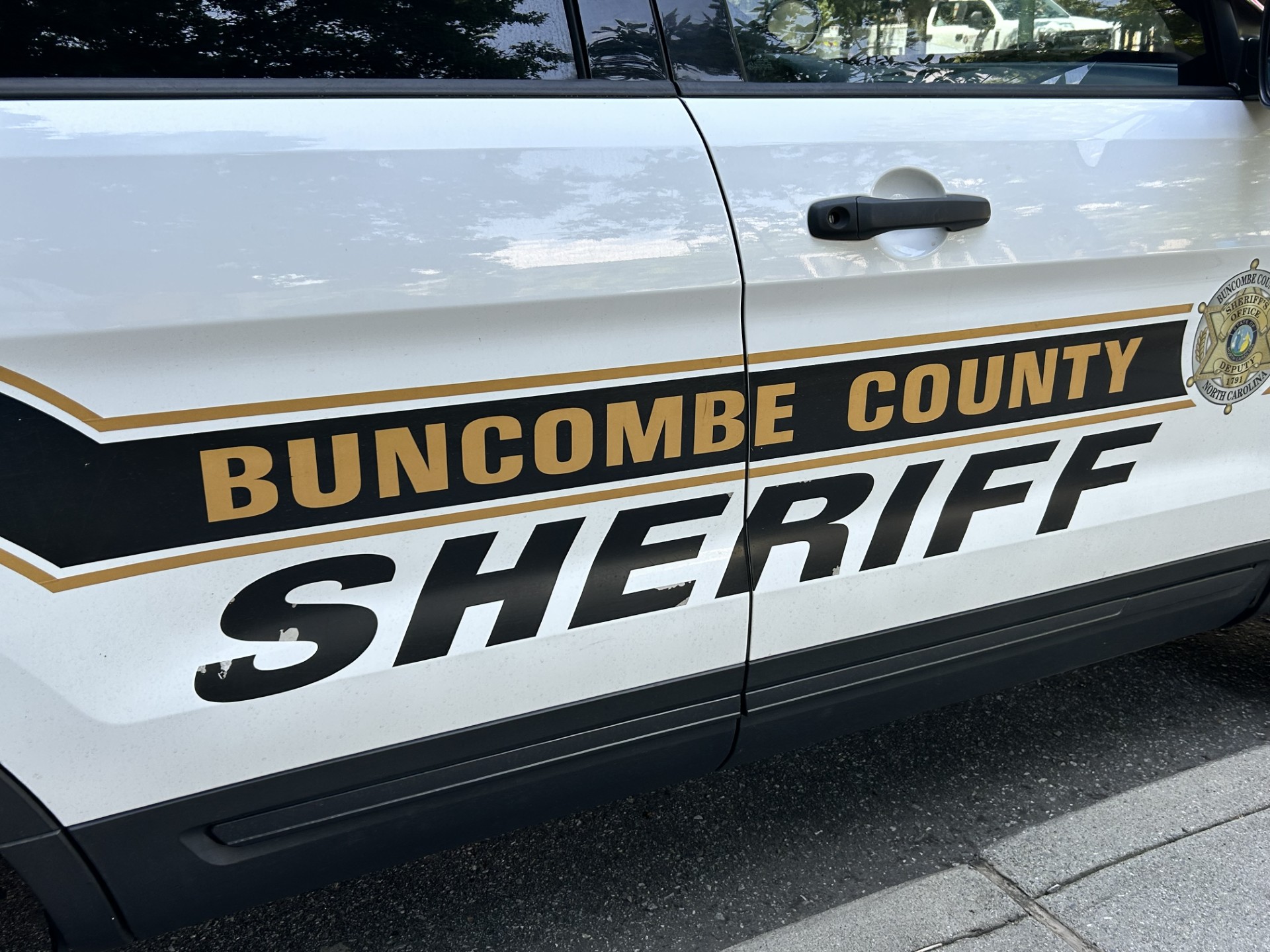 White Buncombe County Sheriff's Office patrol vehicle door with gold and black 'SHERIFF' lettering and sheriff badge visible on the side
