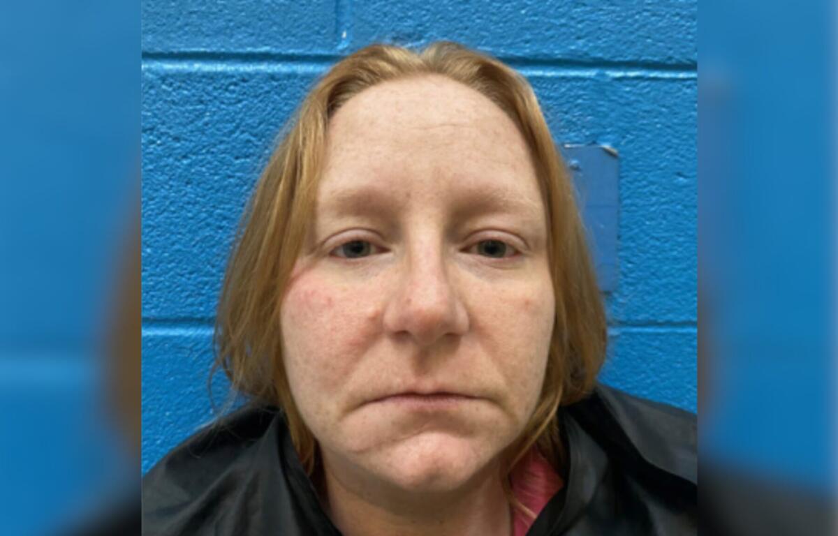 Close-up headshot of a woman with light skin and red-blond hair, standing against a blue wall (mugshot-style photo).
