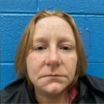 Close-up headshot of a woman with light skin and red-blond hair, standing against a blue wall (mugshot-style photo).