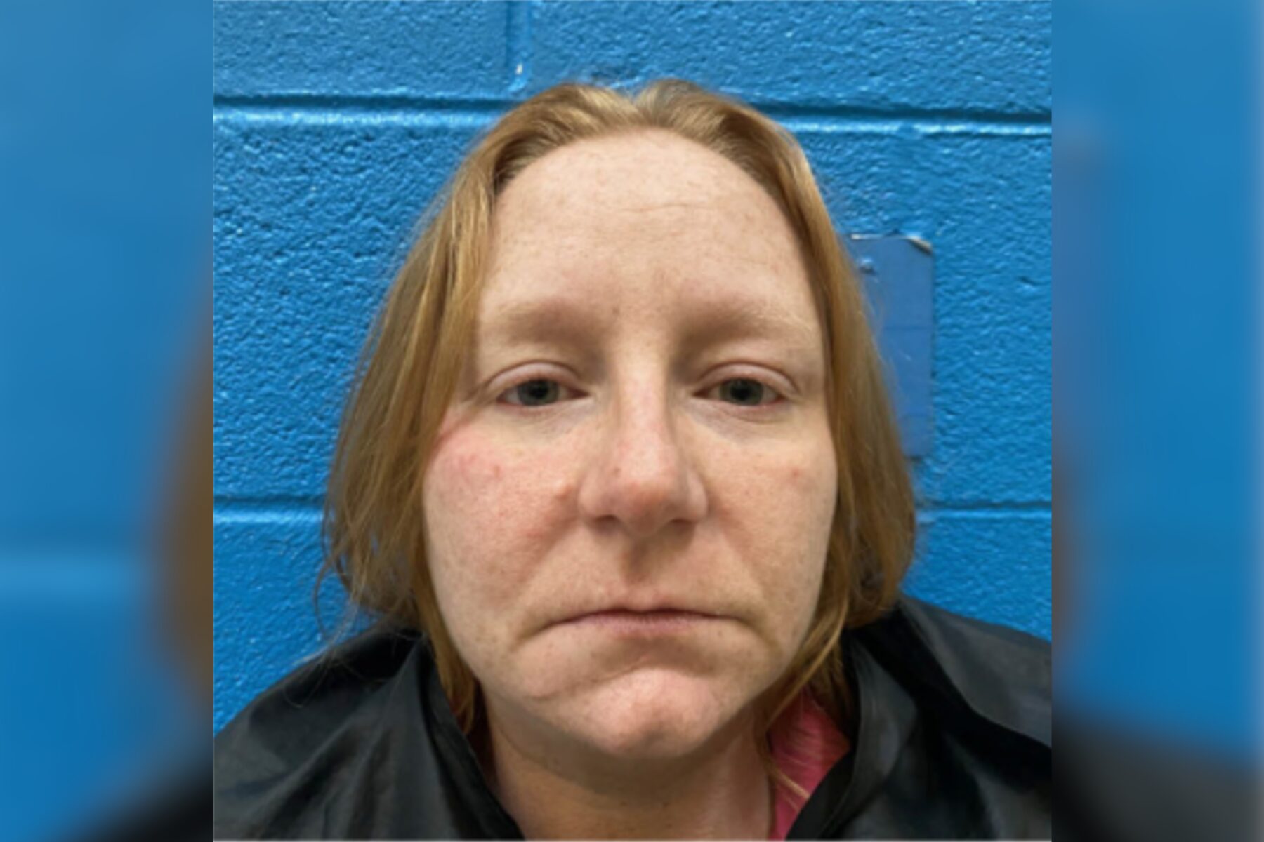 Close-up headshot of a woman with light skin and red-blond hair, standing against a blue wall (mugshot-style photo).