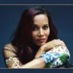 Portrait of a woman with dark hair and magenta highlights, resting her arms and looking at the camera against a blue backdrop, wearing a floral-sleeve top.