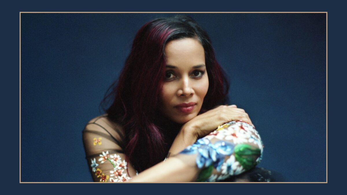 Portrait of a woman with dark hair and magenta highlights, resting her arms and looking at the camera against a blue backdrop, wearing a floral-sleeve top.