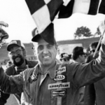 FILE - In this Nov. 8, 1976, file photo, stock car driver David Marcis waves a checkered flag and hold his winner's trophy after the Dixie 500 in Hampton, Ga. Only 39 cars showed up for the second race of the Sprint Cup season, the lowest number of entries in more than two decades. While the reduced numbers won't affect front-runners such as Daytona 500 winner Denny Hamlin _ after all, we're talking about low-budget drivers who probably would've been lapped early in the race _ it does drive home the end of an era. "We've lost a lot," said former driver Dave Marcis, who spent much of his long career simply trying to qualify for races in under-financed cars. (AP Photo/Lennox McLendon, File)