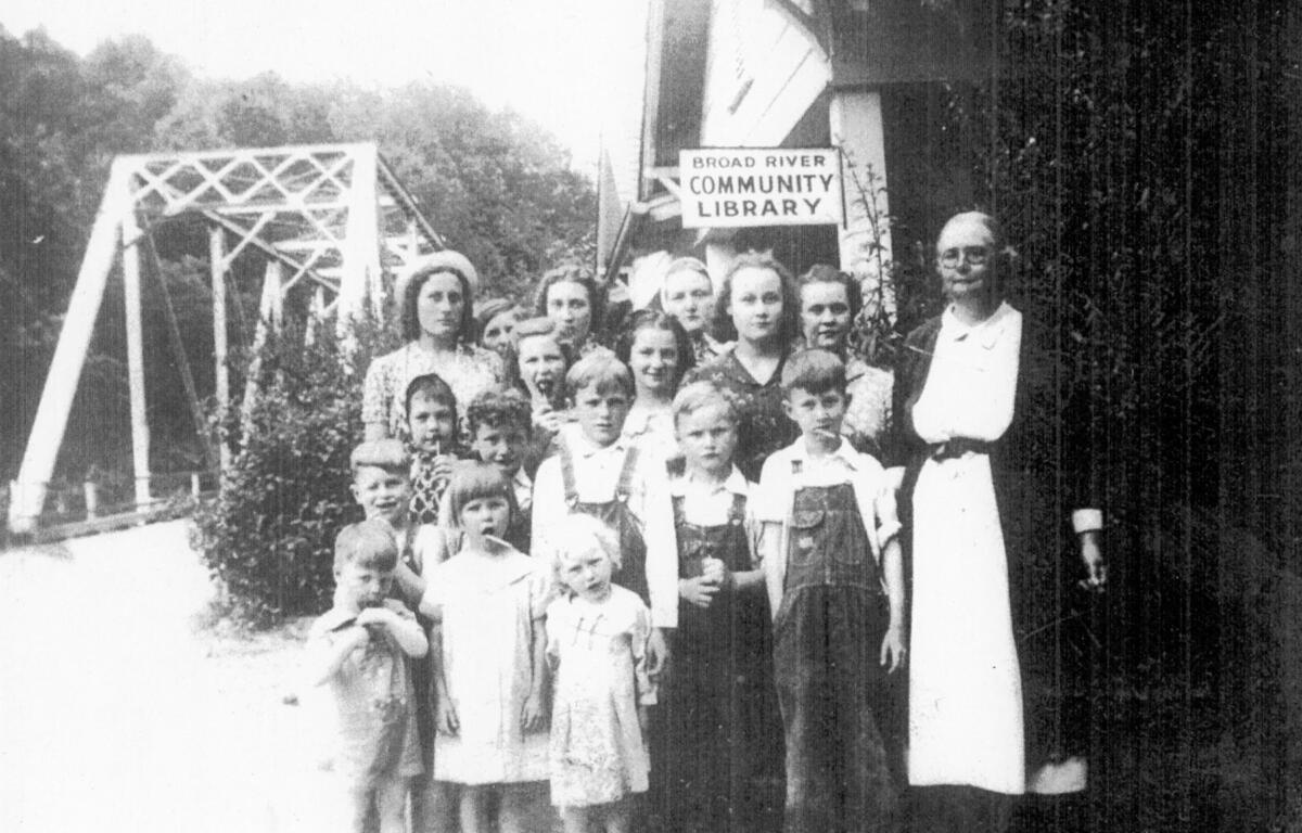 Nora Mae “Aunt Mae” Gilliam stands with children outside the tiny Broad River Community Library, which she operated in rural Buncombe County beginning in 1938. The 12-by-12-foot log building served residents with books and other reading materials.