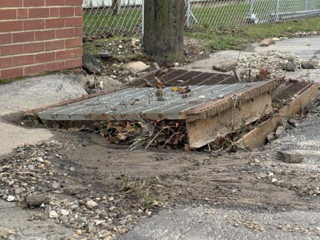 A displaced storm drain grate sits amid gravel, mud and debris along North Avenue in Swannanoa on Monday, Oct. 12, 2026.